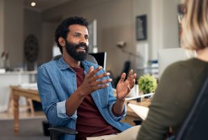 Two casually dressed business individuals in a closed meeting, discussing feedback and planning in a creative office environment. The individual has short black afro hair with a black beard dressed in a maroon tee shirt and a denim long-sleeved on top.