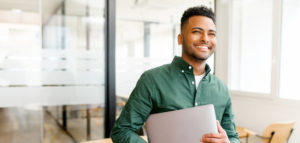 Carefree inspired indian male employee standing in modern office space and holding laptop, cheerful young businessman in green shirt