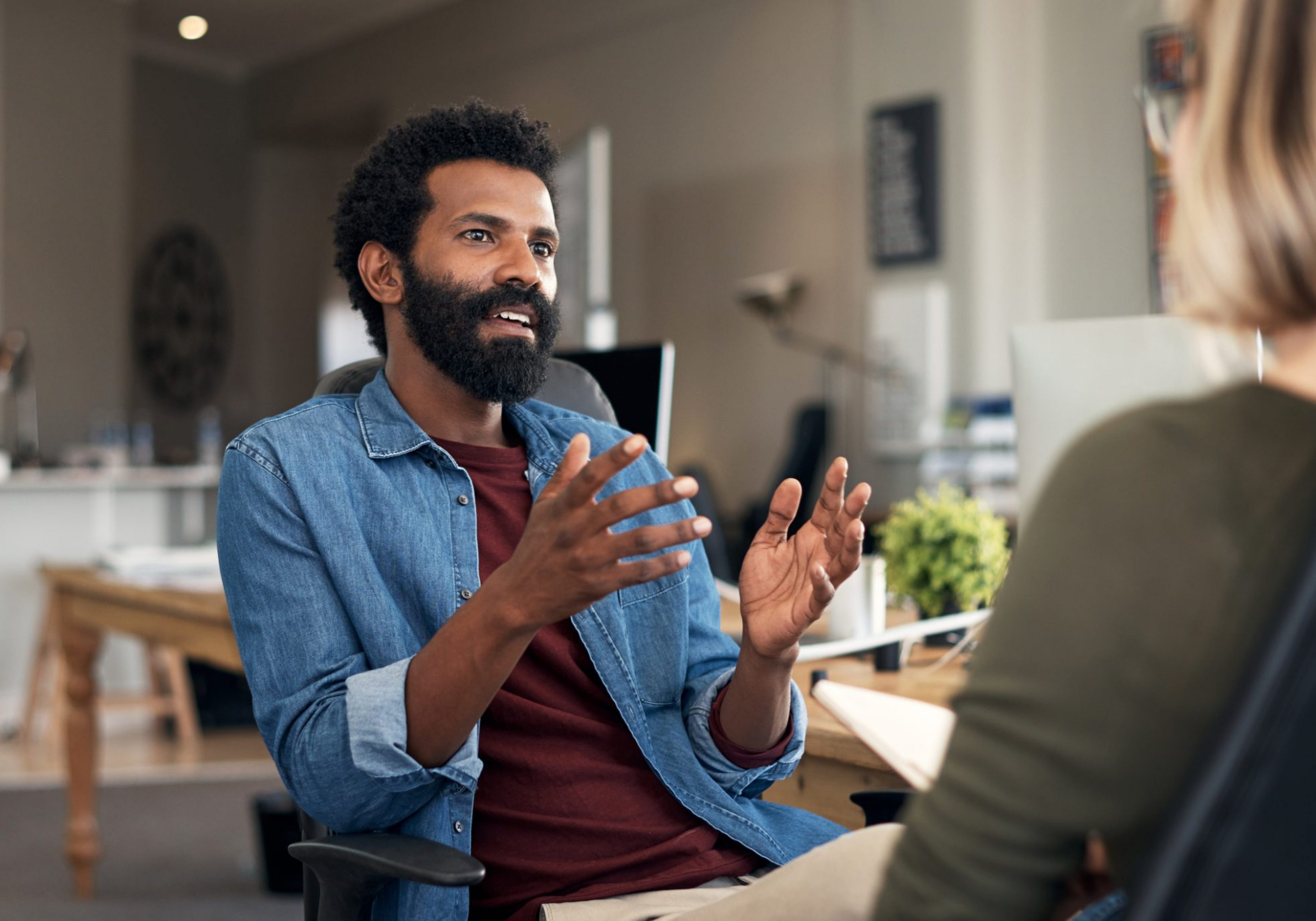 Two casually dressed business individuals in a closed meeting, discussing feedback and planning in a creative office environment. The individual has short black afro hair with a black beard dressed in a maroon tee shirt and a denim long-sleeved on top.