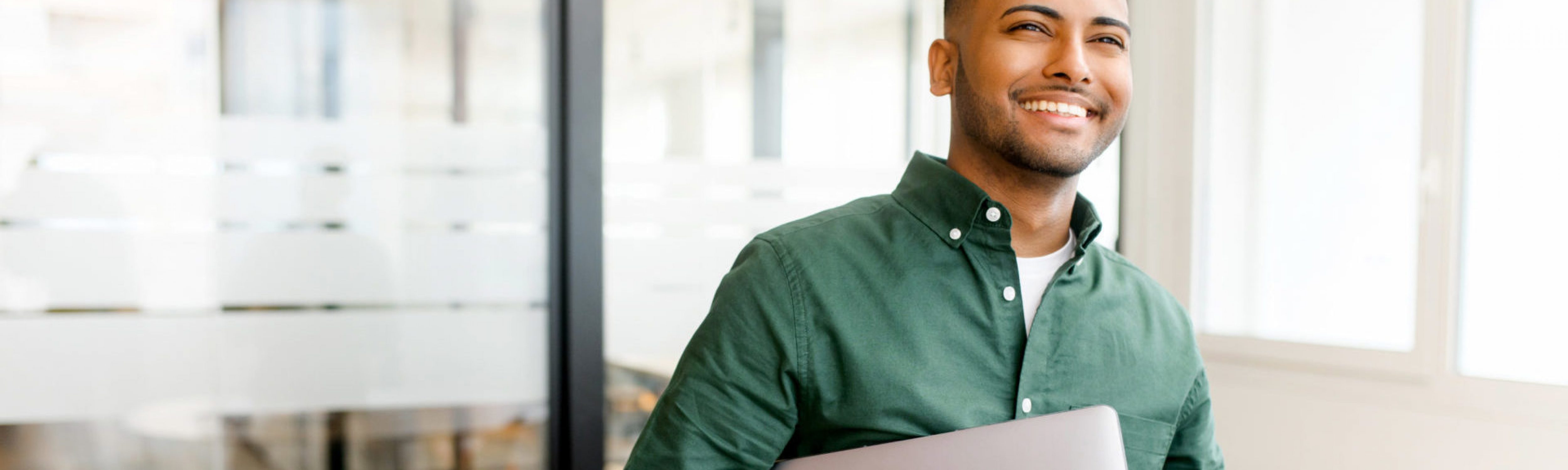 Carefree inspired indian male employee standing in modern office space and holding laptop, cheerful young businessman in green shirt