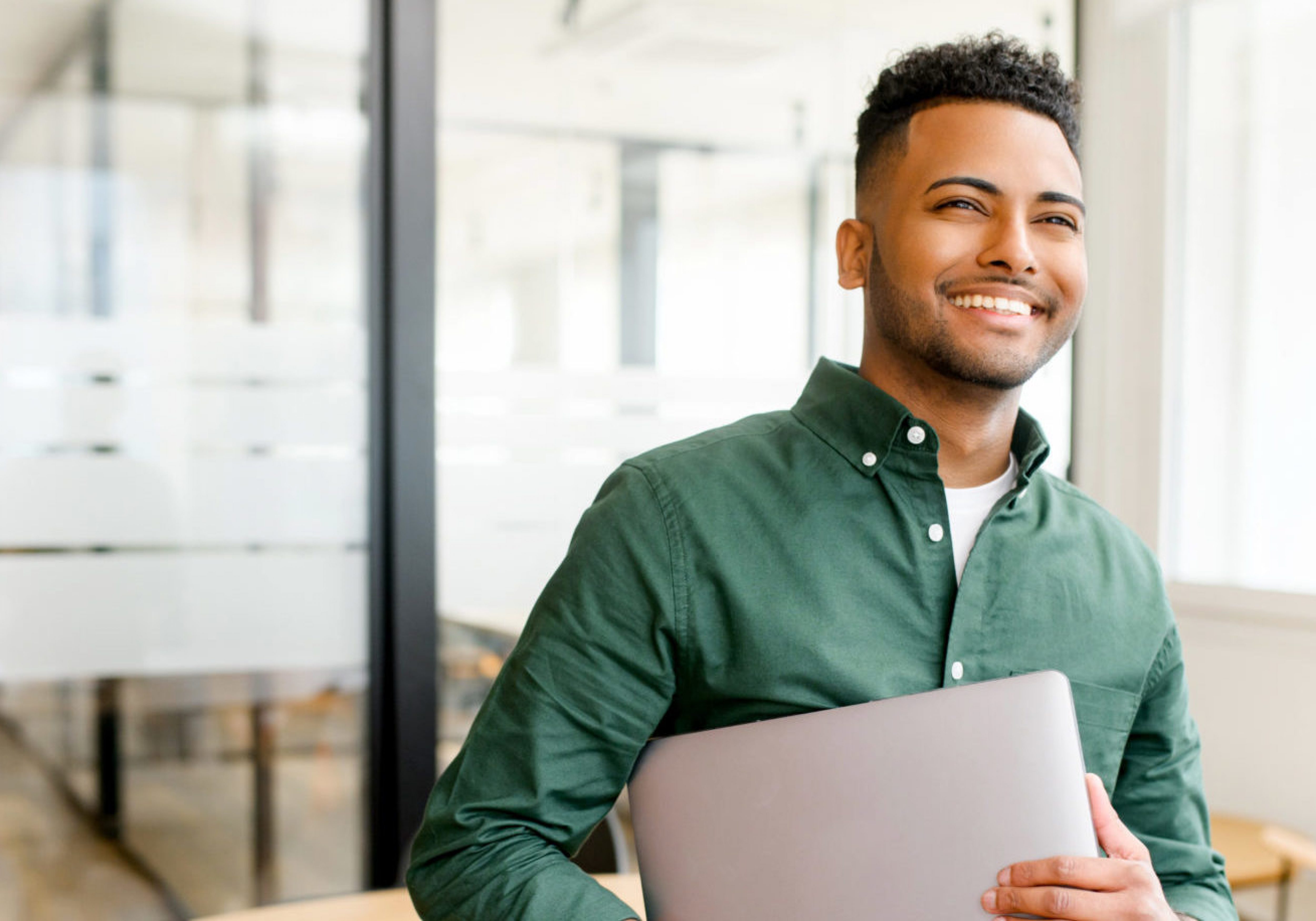 Carefree inspired indian male employee standing in modern office space and holding laptop, cheerful young businessman in green shirt